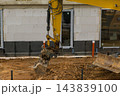 A yellow excavator is actively engaged in digging soil beside a building's foundation at a construction site. 143839100