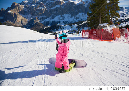 Female snowboarder takes smartphone selfie while kneeling on snowy slope in Dolomites, Italy, with snowboard beside her and dramatic alpine cliffs, resort terrain and bright winter scenery behind 143839671