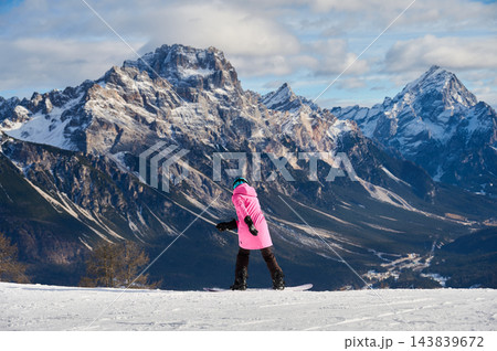 Snowboarder glides across snowy slope in Dolomites, Italy, viewed from behind while riding toward dramatic alpine mountains Punta Sorapis and valley during active winter resort vacation 143839672