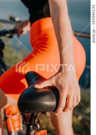 Bicycle, cycling, athlete, woman holding a bike seat while wearing orange sportswear during a sunset ride near a lake shore Bicycle, cycling, athlete, woman holding a bike seat while wearing orange sportswear during a sunset ride near a lake shore 143840135