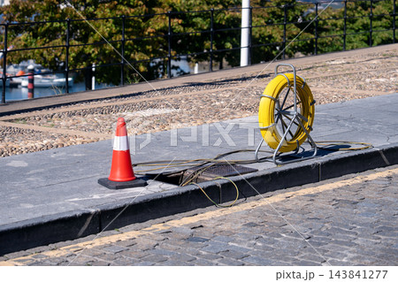 Yellow fiber optic cable reel on a metal stand placed on a concrete sidewalk next to an open utility manhole. Maintenance or telecommunication installation work place Yellow fiber optic cable reel on a metal stand placed on a concrete sidewalk next to an open utility manhole. Maintenance or telecommunication installation work place 143841277