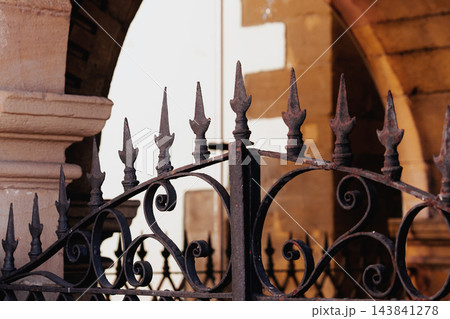 Decorative wrought iron gate spikes with intricate forged scrolls. Vintage rusted metal texture and ancient stone archway architecture in the blurred background Decorative wrought iron gate spikes with intricate forged scrolls. Vintage rusted metal texture and ancient stone archway architecture in the blurred background 143841278