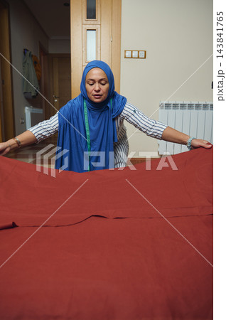 Woman tailor spreads red fabric while preparing handmade clothing in a home sewing workshop, wearing a hijab and measuring tape Woman tailor spreads red fabric while preparing handmade clothing in a home sewing workshop, wearing a hijab and measuring tape 143841765