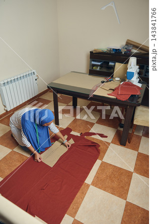 Woman tailors fabric with a paper pattern and measuring tape on the floor in her home sewing workshop beside a sewing machine 143841766
