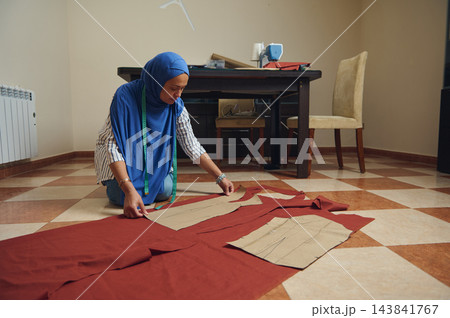 Woman tailors fabric with paper patterns and measuring tape on the floor in a home sewing workshop beside a sewing machine 143841767