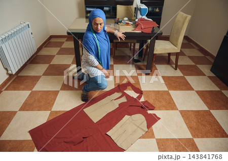 Woman tailor arranges sewing patterns on red fabric for handmade fashion design in a home textile workshop beside a sewing machine Woman tailor arranges sewing patterns on red fabric for handmade fashion design in a home textile workshop beside a sewing machine 143841768