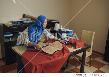 Woman tailor cuts red fabric for handmade clothing patterns with scissors beside a sewing machine in a bright home tailoring workshop 143841769