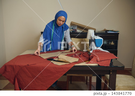 Tailor measures fabric and arranges sewing patterns for handmade clothing in a home fashion workshop beside a sewing machine Tailor measures fabric and arranges sewing patterns for handmade clothing in a home fashion workshop beside a sewing machine 143841770