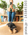 Woman sitting on a shelf, focused on smartphone, with her small cute pet sitting on a rug nearby at home. dog. An authentic moment of life 143845303