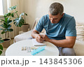 Mature man sits on sofa, noting down medication details in notebook, blister packs and pill organizer are on white side table. Self-care, managing health at home, and adhering to treatment regimens 143845306