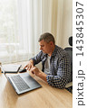 Middle-aged man sits at his home desk, stretching his fingers. He is experiencing wrist discomfort caused by prolonged computer use, repetitive strain injury RSI, and carpal tunnel syndrome 143845307