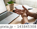 Middle-aged man stretches his wrist next to laptop on wooden desk. Office ergonomics, prevention of repetitive strain injuries, and wrist mobility exercises while working at computer 143845308