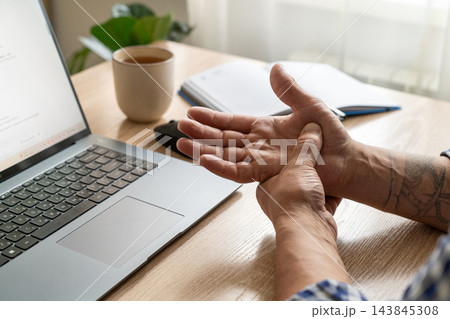 Middle-aged man stretches his wrist next to laptop on wooden desk. Office ergonomics, prevention of repetitive strain injuries, and wrist mobility exercises while working at computer Middle-aged man stretches his wrist next to laptop on wooden desk. Office ergonomics, prevention of repetitive strain injuries, and wrist mobility exercises while working at computer 143845308