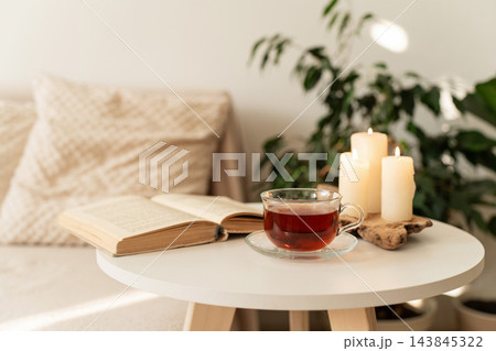 Warm atmosphere creating sense of coziness and relaxation, book, cup of tea, and lit candles on table. Concept of slow living, a mindful lifestyle, cozy home Warm atmosphere creating sense of coziness and relaxation, book, cup of tea, and lit candles on table. Concept of slow living, a mindful lifestyle, cozy home 143845322