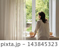 Mature woman in a white blouse stands by large, light-filled window with sheer curtains, gazing at green trees. She feels at peace, experiencing quiet solitude. Authentic slow living 143845323
