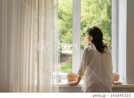 Mature woman in a white blouse stands by large, light-filled window with sheer curtains, gazing at green trees. She feels at peace, experiencing quiet solitude. Authentic slow living 143845323