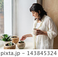 Mature woman stands by bright window holding mug, gazing at small succulent in ceramic pot on windowsill. A moment of domestic life, authenticity 143845324