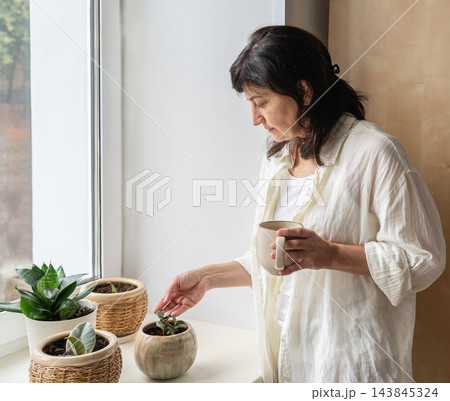 Mature woman stands by bright window holding mug, gazing at small succulent in ceramic pot on windowsill. A moment of domestic life, authenticity 143845324