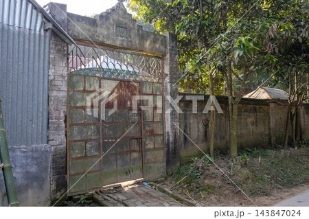 Old rusty iron entrance gate and concrete arch of a rural home. Traditional village house boundary wall surrounded by green trees. 143847024