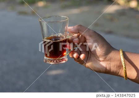 Person holding a glass mug of hot black tea outdoors in natural daylight. Close-up of traditional South Asian chai refreshment from a local stall. 143847025