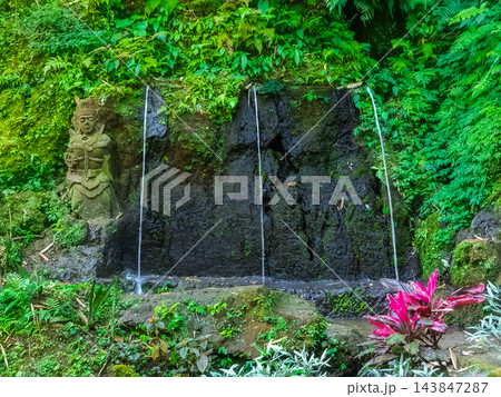 Sacred water spring with stone Hindu guardian statue and water jets on dark volcanic rock wall in tropical jungle, Bali, Indonesia Sacred water spring with stone Hindu guardian statue and water jets on dark volcanic rock wall in tropical jungle, Bali, Indonesia 143847287