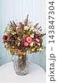Dried wildflower bouquet with strawflowers and eucalyptus in crystal vase on white table against light blue wall 143847304