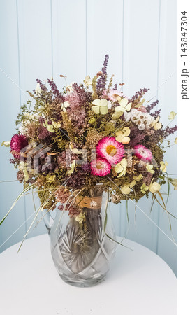 Dried wildflower bouquet with strawflowers and eucalyptus in crystal vase on white table against light blue wall Dried wildflower bouquet with strawflowers and eucalyptus in crystal vase on white table against light blue wall 143847304