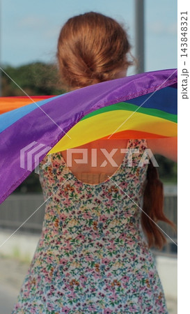 Vertical video. Back view of young Woman holding LGBT rainbow flag while walking on street on a sunny day. 143848321