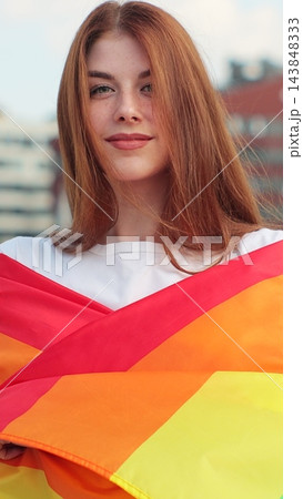 Vertical video. Portrait of a young woman with long red hair covering herself in lgbt flag while looking to camera. 143848333