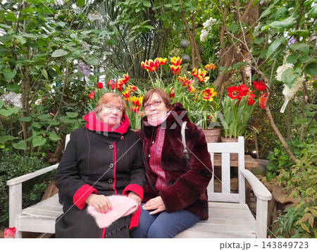Two older women sit together on a white bench among flowering plants, enjoying companionship during a visit to a lush greenhouse Two older women sit together on a white bench among flowering plants, enjoying companionship during a visit to a lush greenhouse 143849723