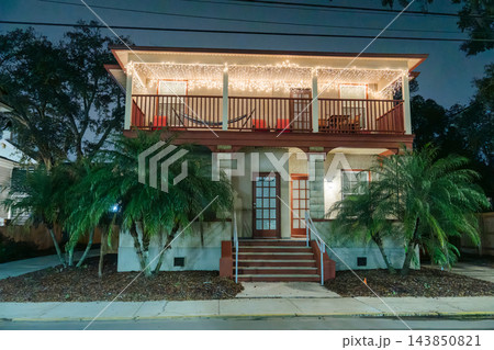 White Christmas lights illuminate the upper balcony and roof of a two-story house. Palm trees, red chairs, and a hammock create a tropical holiday scene. 143850821