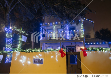 Blue and white Christmas lights illuminate a two-story house, balcony, fence, and large tree. Red bows and green garland decorate the entrance on a festive Florida evening. Blue and white Christmas lights illuminate a two-story house, balcony, fence, and large tree. Red bows and green garland decorate the entrance on a festive Florida evening. 143850826