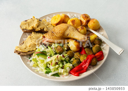 A plate holds baked chicken leg, potatoes, fresh salad, roasted Brussels sprouts, and chickpea bread. This meal is ready to eat at a dining table A plate holds baked chicken leg, potatoes, fresh salad, roasted Brussels sprouts, and chickpea bread. This meal is ready to eat at a dining table 143850933