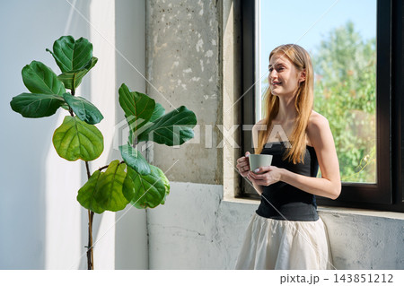 Portrait of smiling dreamy young woman with cup, profile view near window 143851212