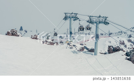 Cable car gondolas suspended above snowy alpine slopes with rocky outcrops and utility towers Cable car gondolas suspended above snowy alpine slopes with rocky outcrops and utility towers 143851638