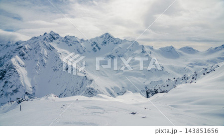 Snow-covered Caucasus mountain ridges and ski slope under cloudy winter sky with distant peaks 143851656