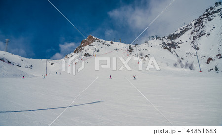 Skiers descending groomed snowy slope below rocky mountain peaks under blue sky and chairlifts 143851683