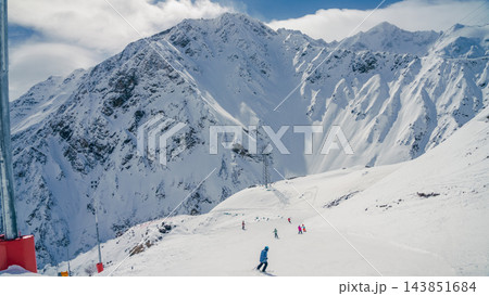 Skiers descending sunlit snowy slope below rugged Mount Elbrus peaks in Caucasus panorama 143851684