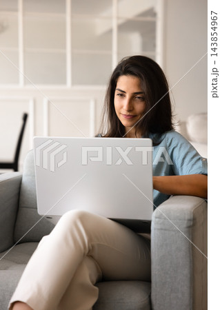 Beautiful millennial girl sitting in comfy armchair using modern notebook Beautiful millennial girl sitting in comfy armchair using modern notebook 143854967