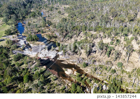 Aerial photograph of a shallow river flowing over broad rock ledges into pools, surrounded by native woodland and rugged bushland. 143857244