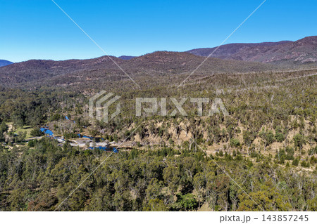 Aerial photograph of native forest and rolling mountain ridges surrounding a winding river in a remote rural valley beneath a blue sky. 143857245