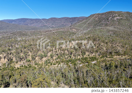 Aerial photograph of rugged forested hills and rocky ridgelines rising above dry native bushland beneath a clear blue sky. 143857246