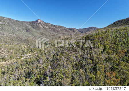 Aerial photograph of a rocky outcrop rising above forested hills and native eucalypt woodland beneath a vivid blue sky. Aerial photograph of a rocky outcrop rising above forested hills and native eucalypt woodland beneath a vivid blue sky. 143857247