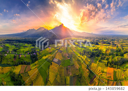 Aerial view of golden sunlight illuminating agricultural fields and rural landscapes beneath dramatic clouds, with mountain silhouettes enhancing the scenic countryside panorama. Aerial view of golden sunlight illuminating agricultural fields and rural landscapes beneath dramatic clouds, with mountain silhouettes enhancing the scenic countryside panorama. 143859682