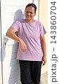 Asian Woman rests against white entry door with furrowed, thoughtful expression in direct sunlight. Adult woman wears lavender T shirt and black trousers, one arm relaxed near door handle. 143860704