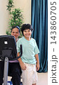 Happy child poses beside seated woman and medical scale in clinic. Smiling woman sits behind weighing equipment while child stands in mint shirt near blue curtains and leafy plant. 143860705
