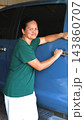 Woman poses beside blue car with hand on its chrome door handle and arm extended across its side. Woman meets viewers gaze with relaxed expression, dark windows behind her, standing close to car door. 143860707