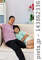 Happy woman stands beside smiling boy outdoors, with her arm around him. Affectionate pair pose beneath large palm fronds, creating friendly family portrait in leafy setting. Both look toward camera. 143862336