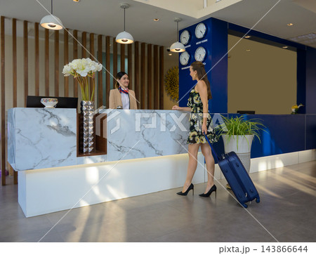 Modern hotel lobby with marble reception desk. Female guest checking in with suitcase while uniformed receptionist assists. 143866644