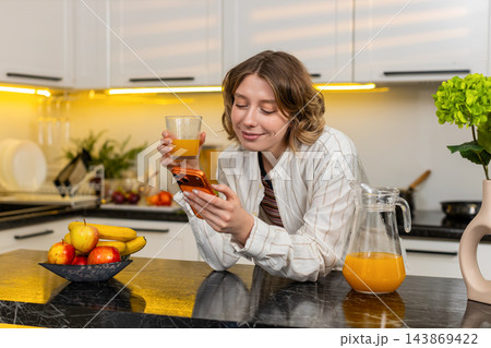 Young woman using smartphone while drinking juice in home kitchen during healthy breakfast routine 143869422